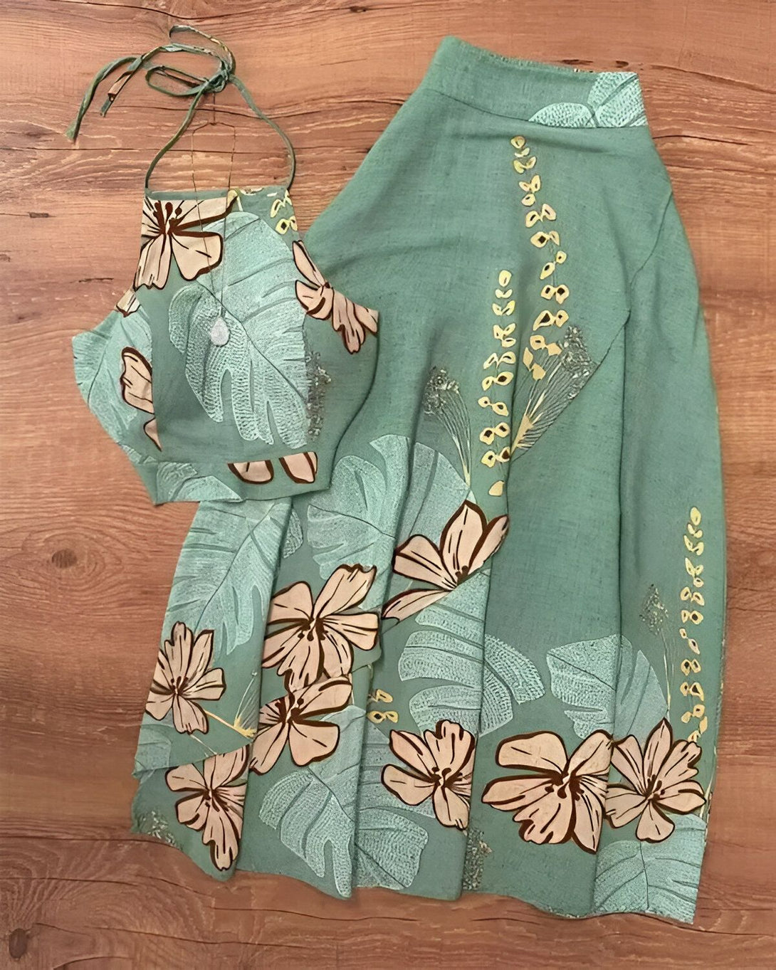 Une image d'un ensemble de vêtements pour femme de couleur vert clair avec un motif de fleurs et de feuilles tropicales. L'ensemble est composé d'un haut court et d'une jupe longue. Le haut est de type bandeau et possède des bretelles fines. La jupe est taille haute et possède une fente sur le côté. L'ensemble est fabriqué dans un tissu léger et fluide.