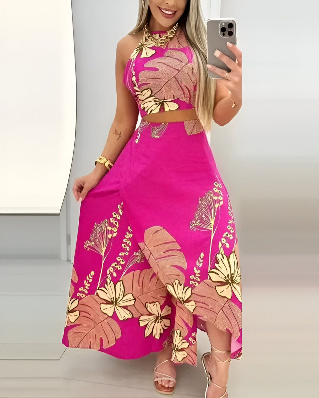 Une femme porte un ensemble deux pièces rose avec un motif de feuilles tropicales. Le haut est un crop top à col montant et la jupe est longue et fendue sur le côté. La femme est souriante et porte un collier doré. Elle est debout dans un miroir et tient son téléphone portable à la main.
