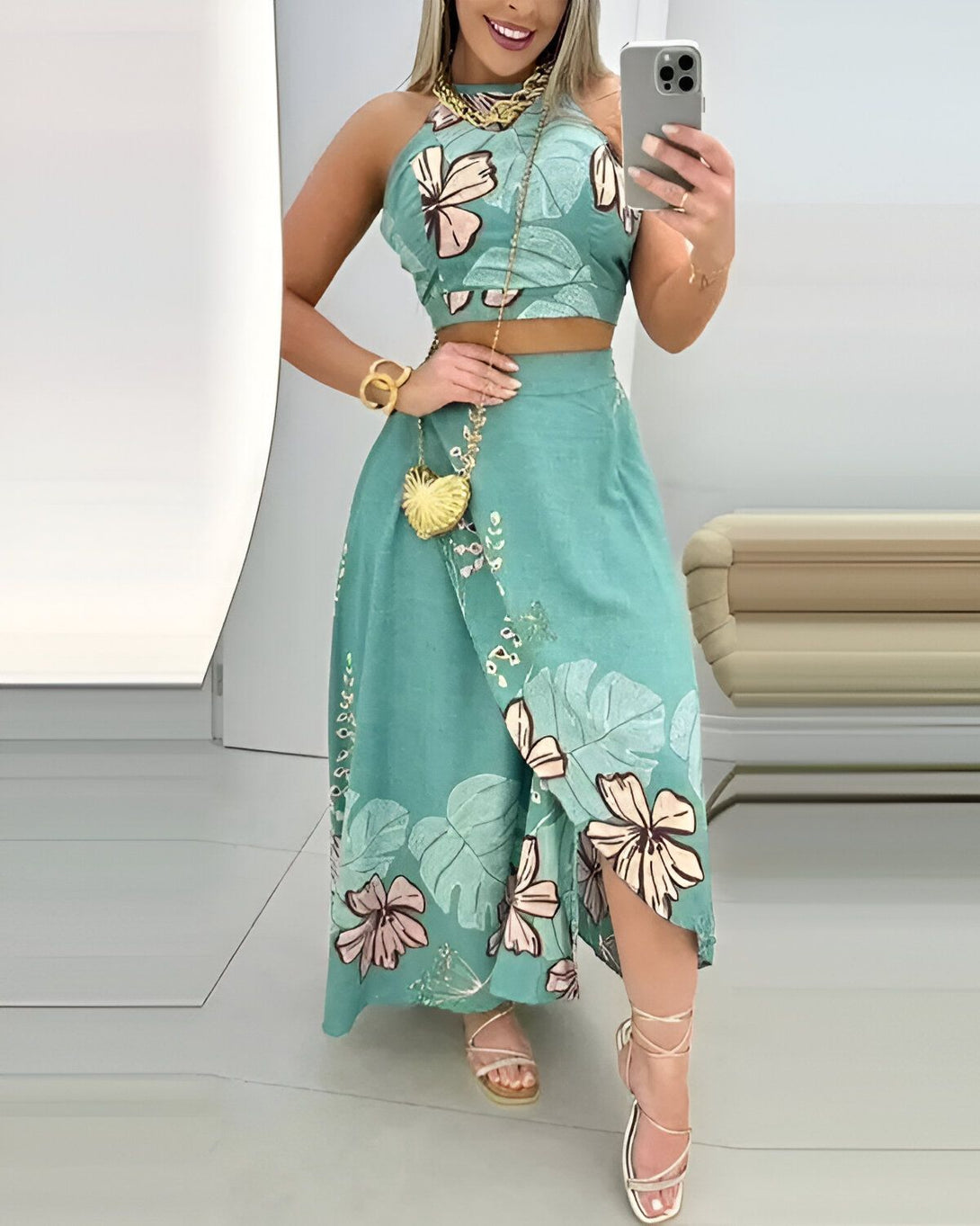 Une femme porte un ensemble deux pièces vert à motifs fleuris. Le haut est un crop top et la jupe est longue et fendue sur le côté. Elle porte des sandales à talons hauts et un collier doré.