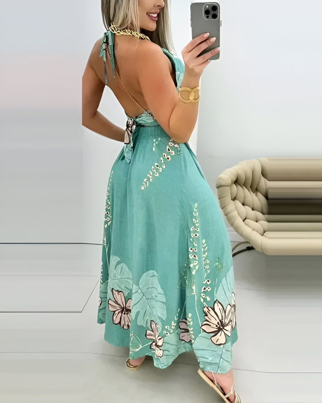 Une femme porte une robe longue verte à motifs de fleurs tropicales. La robe est dos nu et possède un lien à nouer au niveau du cou ainsi qu'au niveau de la taille.