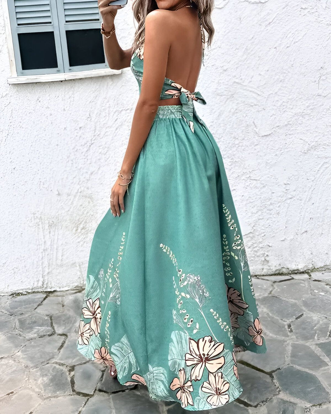 Une femme porte une robe longue vert émeraude à motifs fleuris. La robe est dos nu et possède un lien à nouer au niveau du cou.