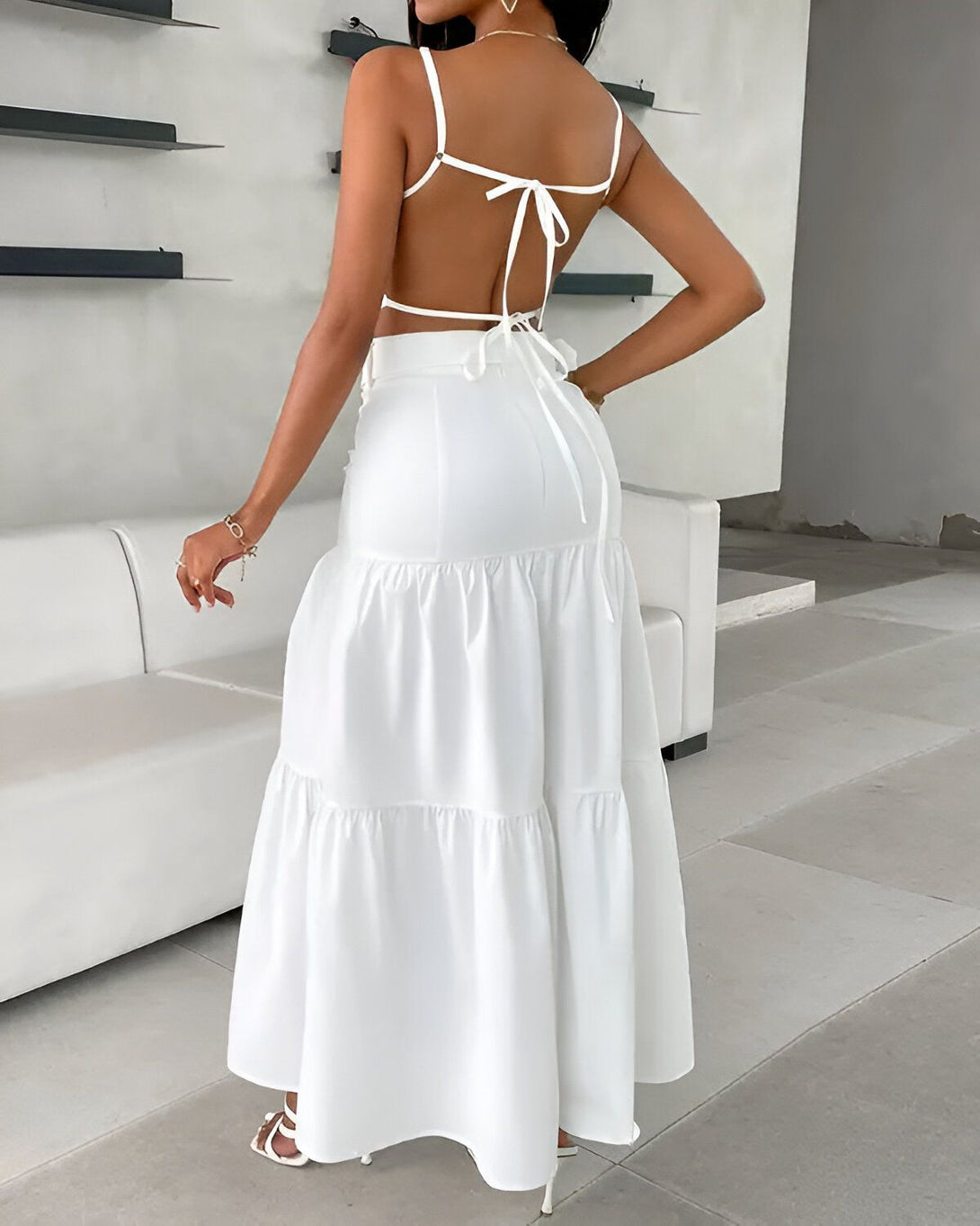 Une femme porte une longue robe blanche dos nu. La robe est fendue sur le côté et possède une traîne. Elle est accessoirisée d'un collier et de sandales blanches.