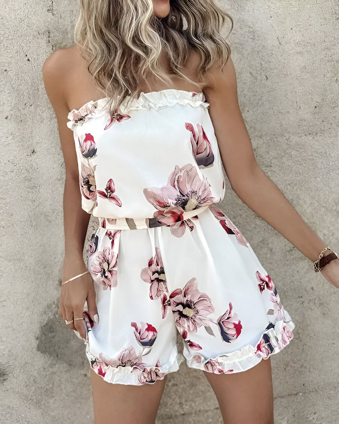Une jeune femme porte une combinaison short blanche à motifs floraux roses. Elle est dos nu et a les cheveux longs blonds.