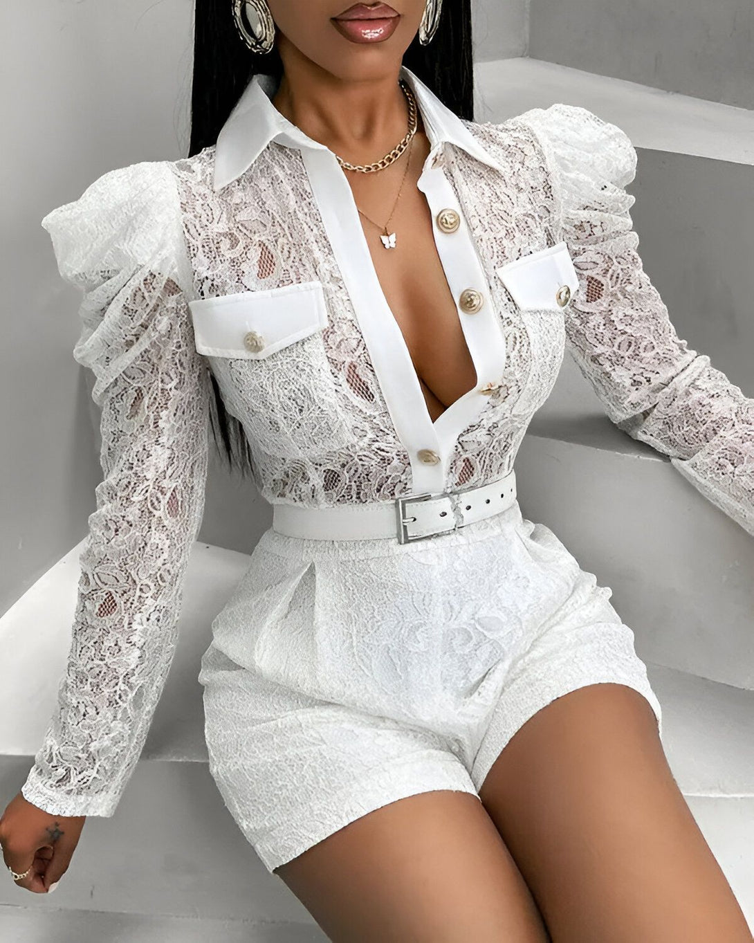 Une femme porte une combinaison short blanche en dentelle. La combinaison est composée d'un haut à manches longues et d'un short taille haute. Le haut est boutonné sur le devant et possède des épaulettes bouffantes. Le short est ceinturé à la taille. La femme porte également un collier et des boucles d'oreilles.