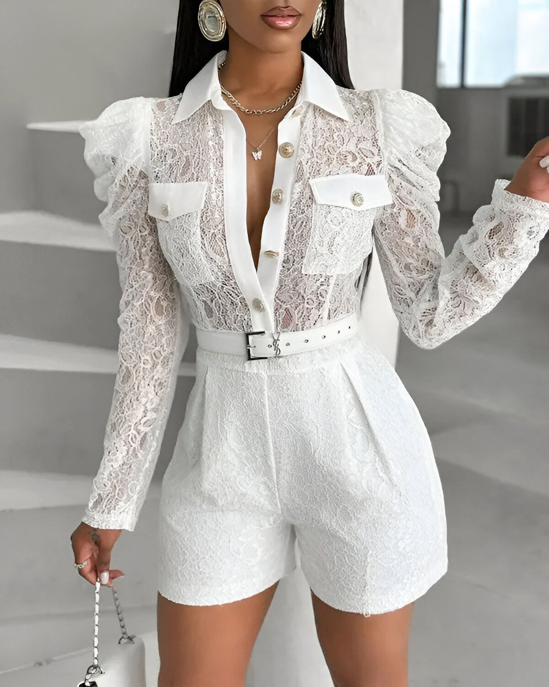 Une femme porte une combinaison blanche en dentelle. La combinaison est composée d'un haut à manches longues et d'un short. Le haut est boutonné et possède un col. Le short est taille haute et possède une ceinture. La combinaison est doublée.