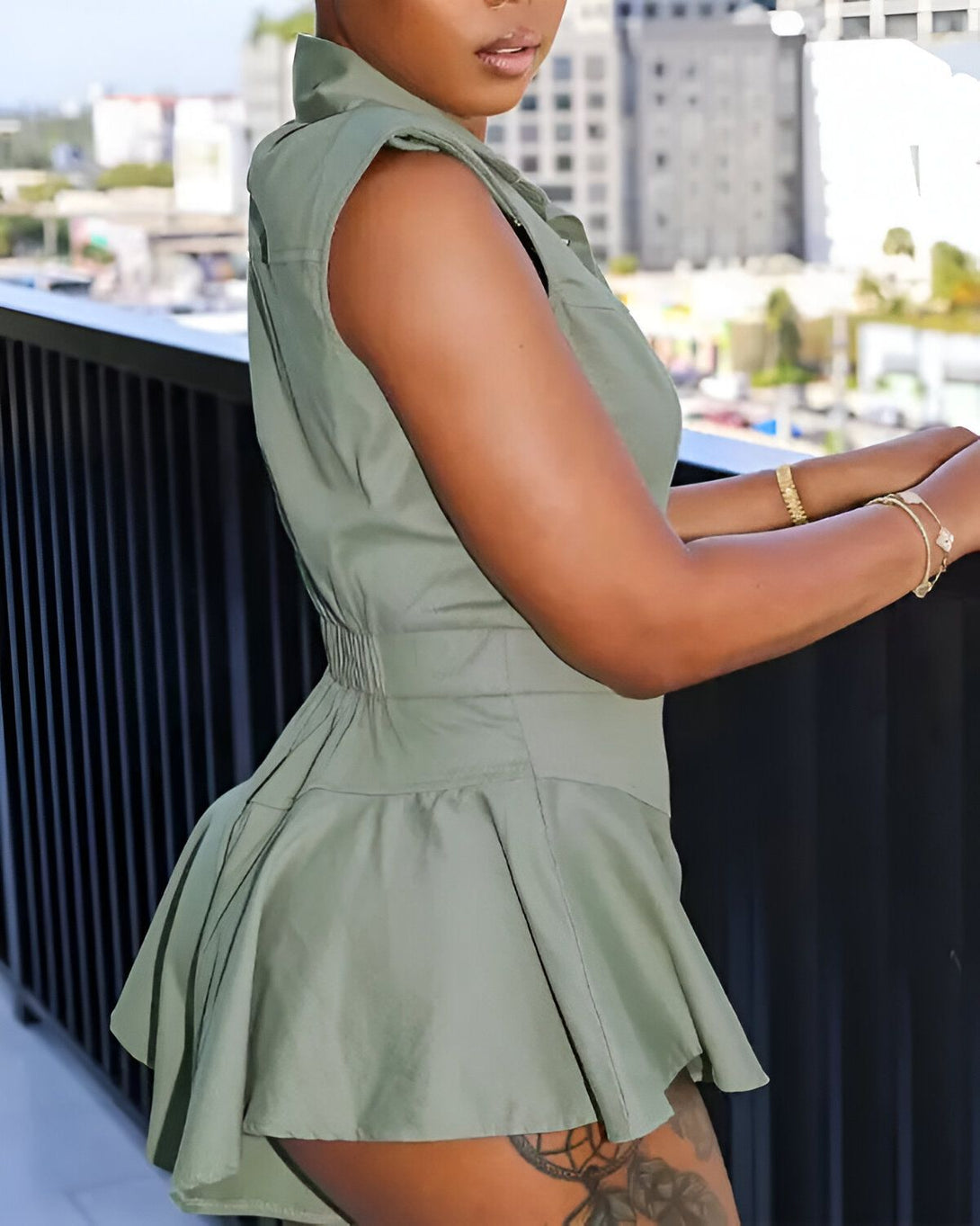 Une femme porte une robe verte courte à manches courtes et à volants. Elle est debout sur un balcon et regarde la ville.