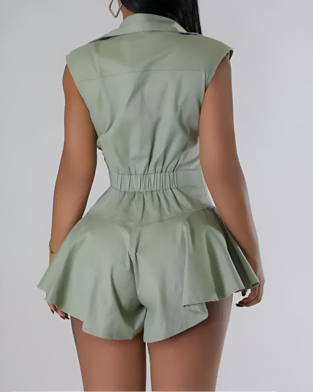 Une femme porte une combinaison short verte dos nu. La combinaison est cintrée à la taille et évasée au niveau des hanches. Elle est également dotée de poches latérales.