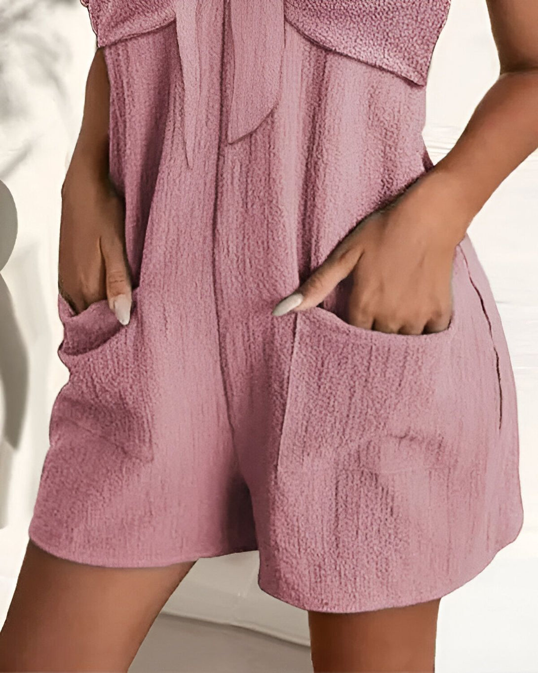 Une femme porte une combinaison short rose avec un nœud sur le devant. Elle a les mains dans les poches.