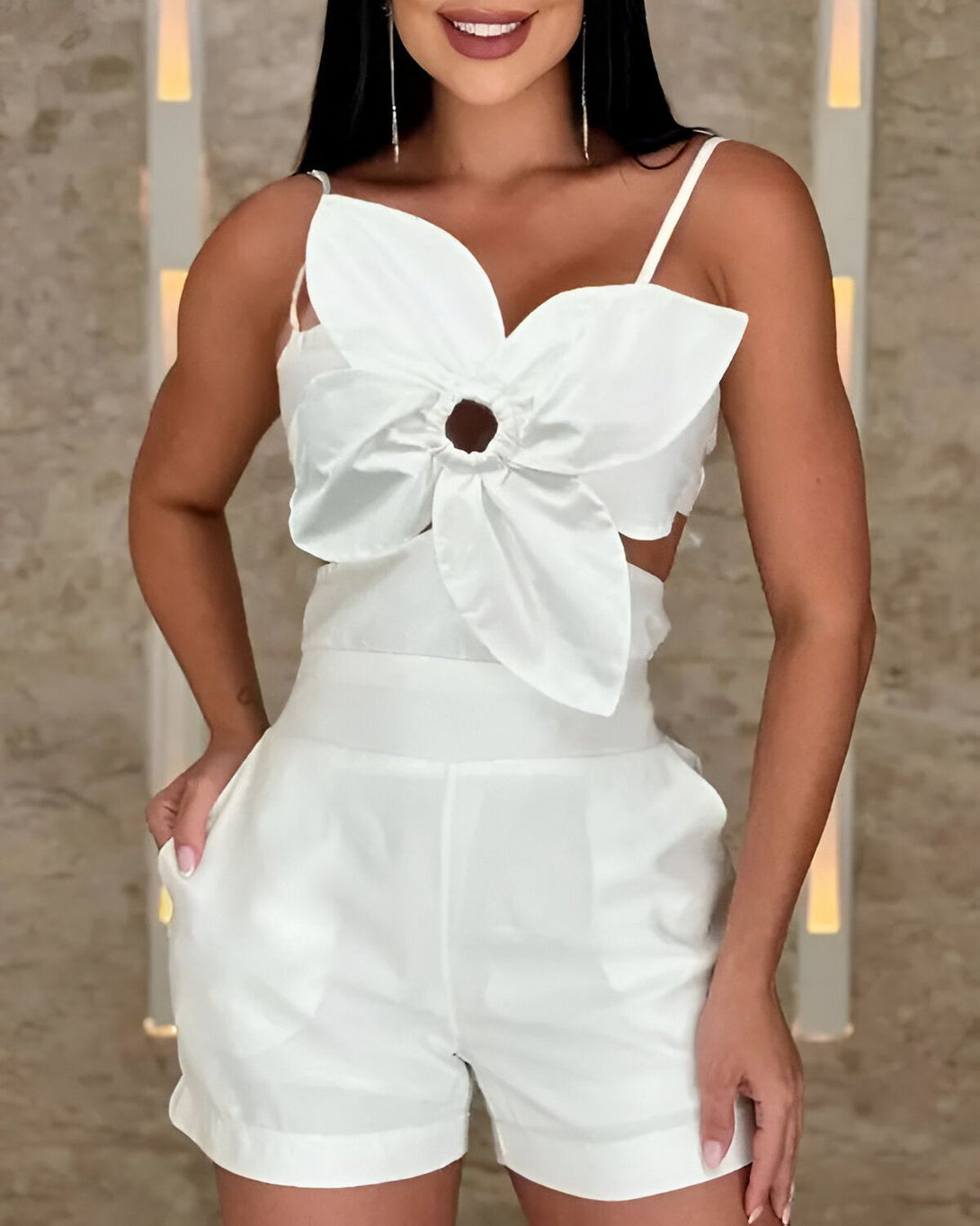Une jeune femme porte une combinaison short blanche. La combinaison est composée d'un haut à bretelles avec un détail en forme de fleur au niveau de la poitrine et d'un short taille haute. La femme est souriante et a les cheveux longs noirs. Elle porte des boucles d'oreilles et un bracelet. Elle est debout dans une pièce avec un mur en pierre et des lumières.