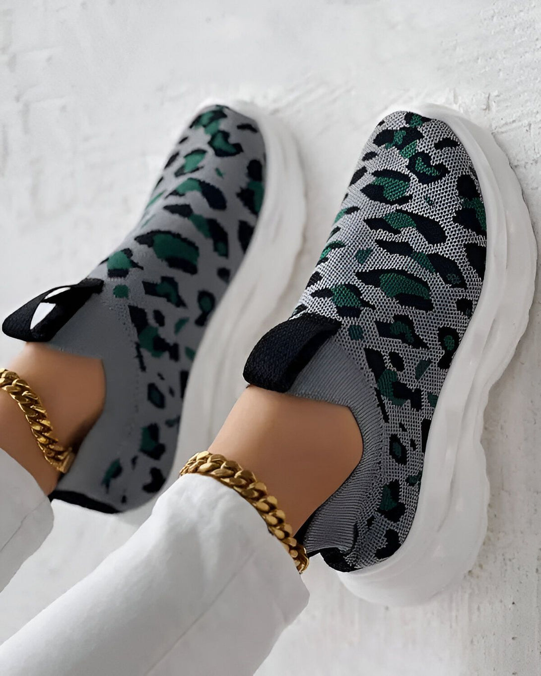 Une femme porte des baskets grises et vertes à motif léopard. Les baskets sont enfilées et ont une semelle blanche. La femme porte également un pantalon blanc et un bracelet doré à la cheville.