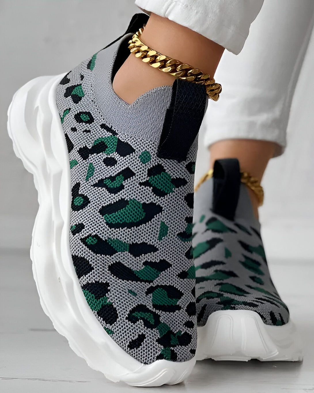 Une femme porte des baskets grises et vertes à motif léopard. Les baskets sont enfilées et ont une semelle blanche. La femme porte également un pantalon blanc et une cheville dorée.