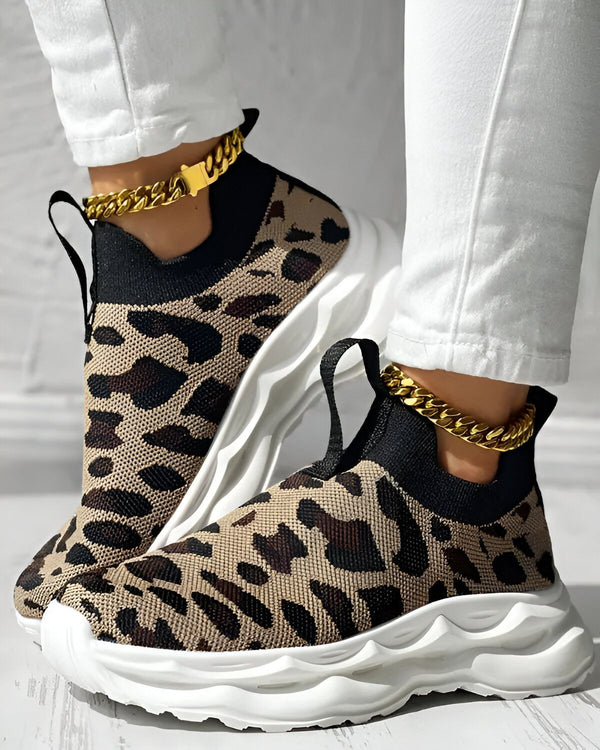 Une femme porte des baskets léopard avec une chaîne dorée autour de la cheville. Elle porte également un jean blanc et un haut blanc. Les baskets sont en maille et ont une semelle blanche. La femme est debout sur un sol en bois.