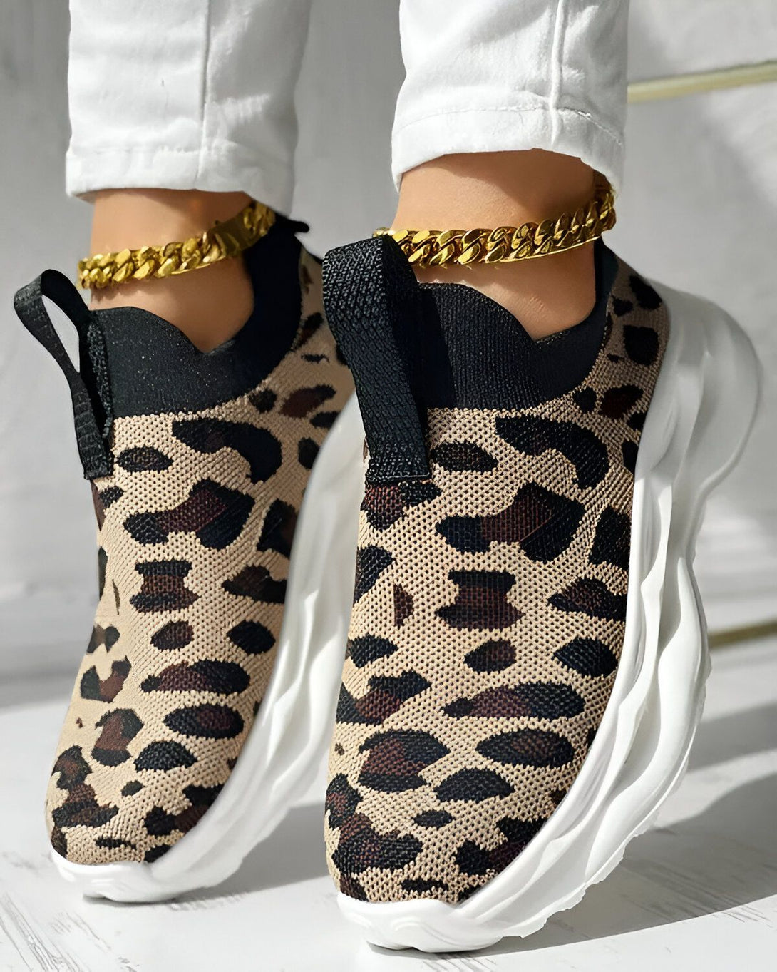 Une femme porte des baskets léopard avec une semelle blanche. Elle porte également un pantalon blanc et des chaussettes noires.