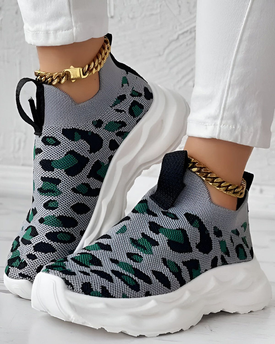 Une femme porte des baskets grises et vertes à motif léopard. Les baskets sont enfilées et ont une semelle blanche. La femme porte également un jean blanc et une chaîne dorée à la cheville.