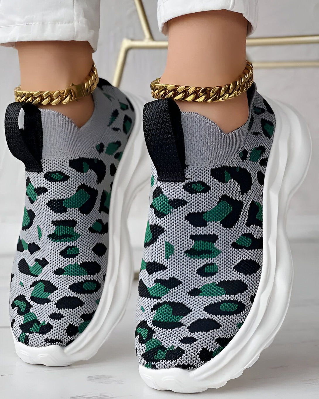 Une femme porte des baskets grises et vertes à motif léopard. Les baskets sont en maille et ont une semelle blanche. La femme porte également un pantalon blanc et un chemisier noir. Elle est debout sur un sol en marbre.