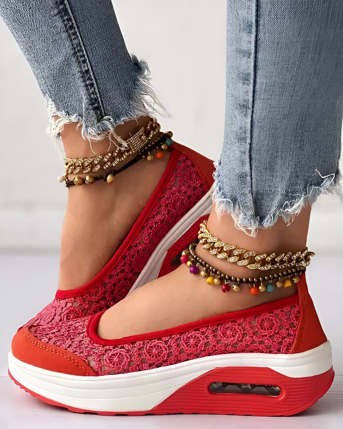 Une femme porte des chaussures rouges en dentelle avec des semelles blanches. Elle porte également des jeans bleus déchirés et des bracelets de cheville.