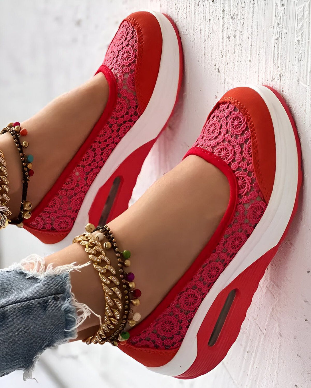 Une femme porte des baskets rouges en dentelle avec une semelle blanche. Elle porte également des bracelets de cheville en perles et en or.