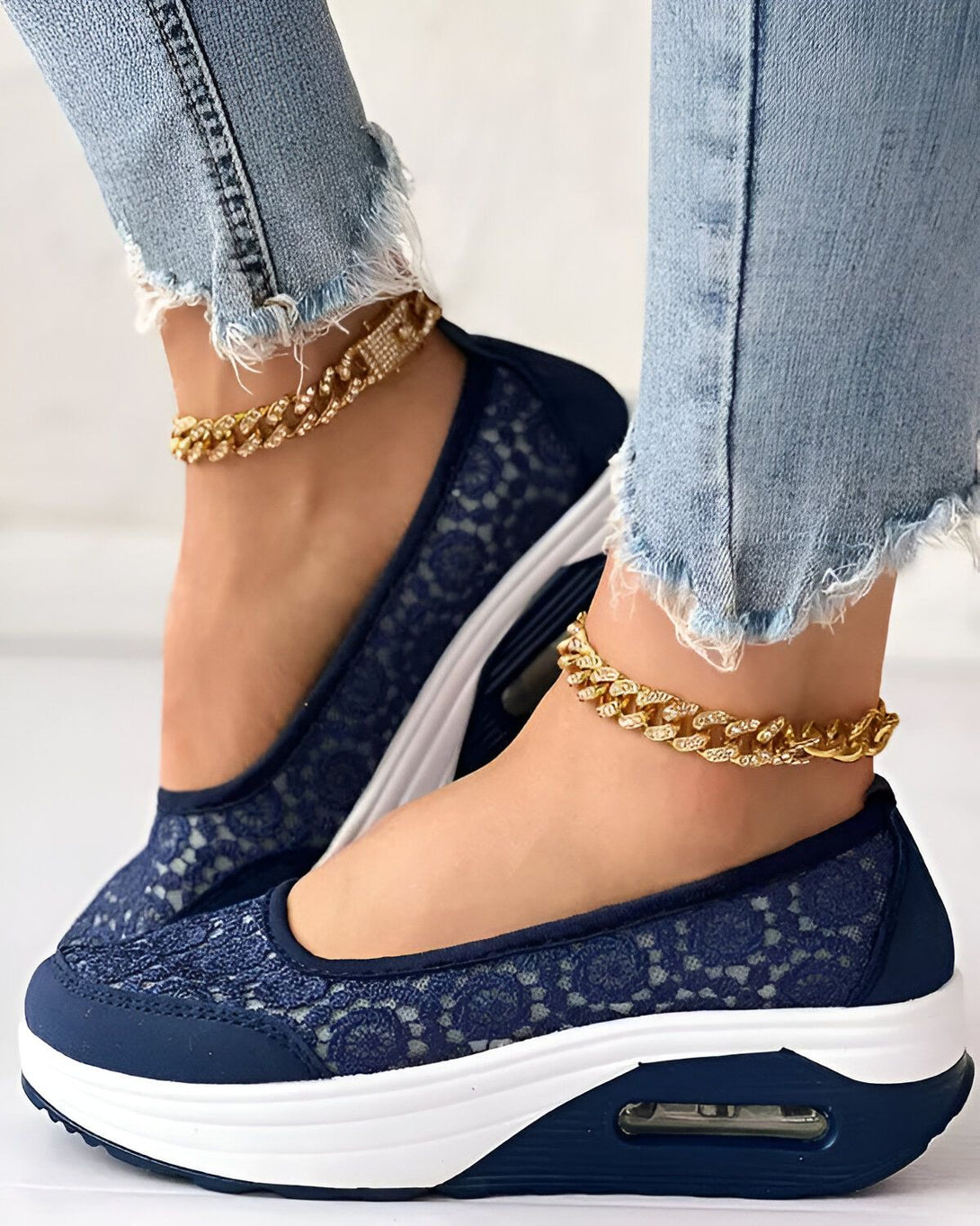 Une femme porte des chaussures bleues en dentelle avec une semelle blanche. Elle porte également un jean bleu et des bracelets de cheville en or.