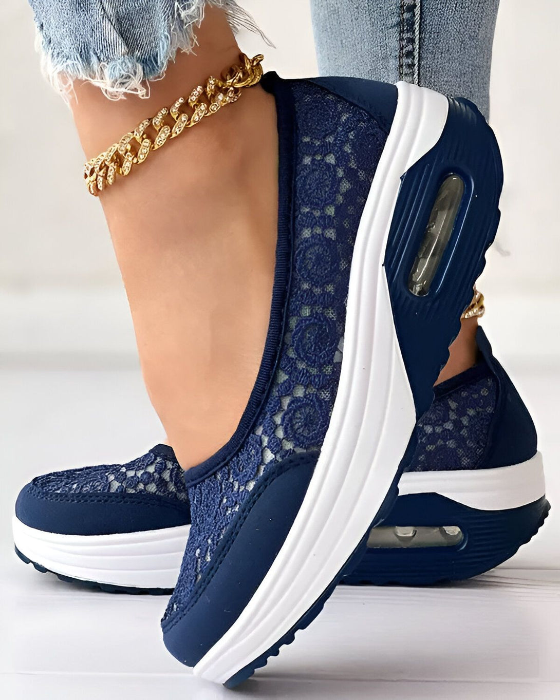 Une femme porte des baskets bleues en dentelle avec une semelle blanche. Elle porte également un jean et un bracelet de cheville doré.