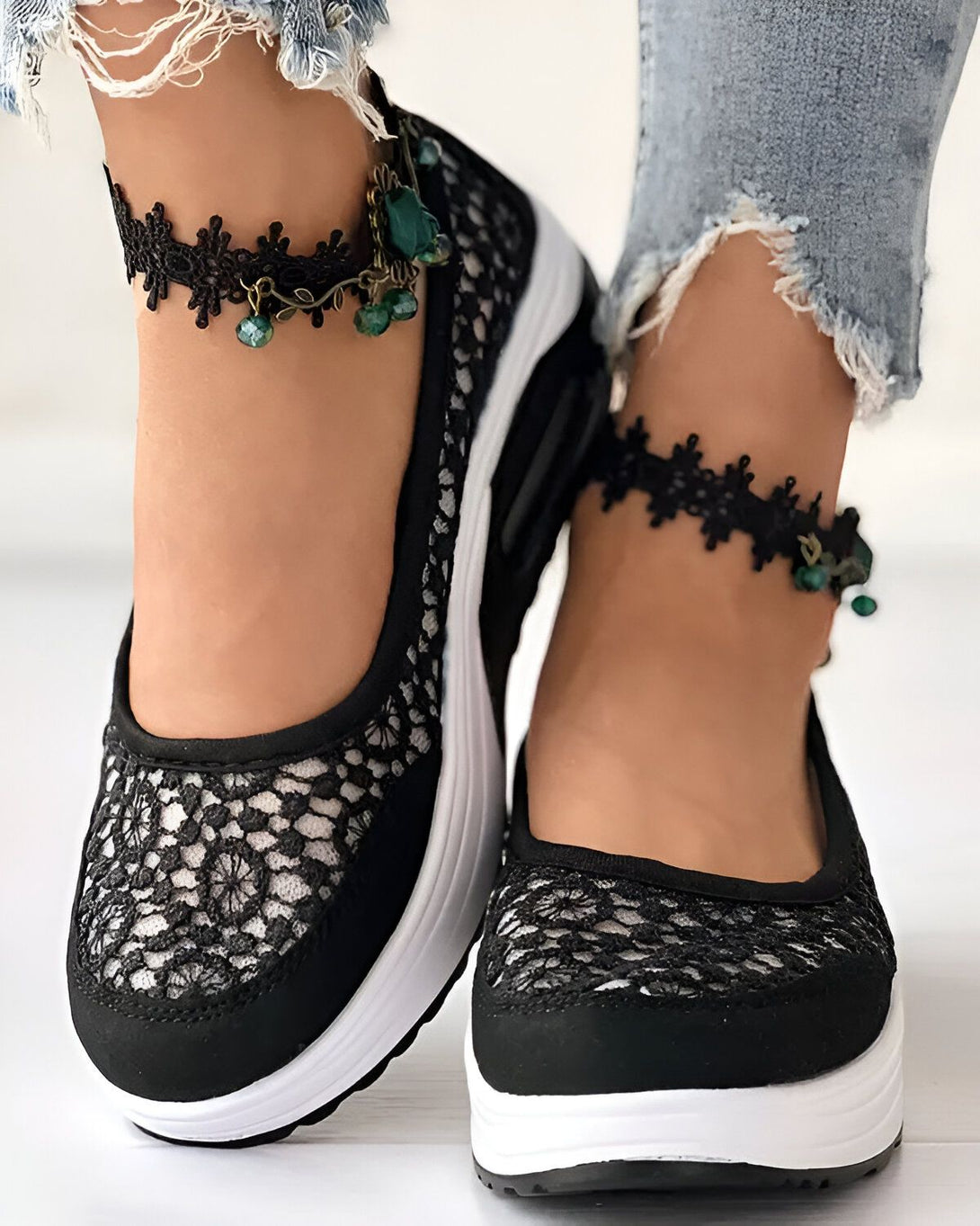 Une femme porte des chaussures noires en dentelle avec des semelles blanches. Elle porte également des bracelets de cheville en dentelle noire avec des breloques en forme de feuilles vertes.