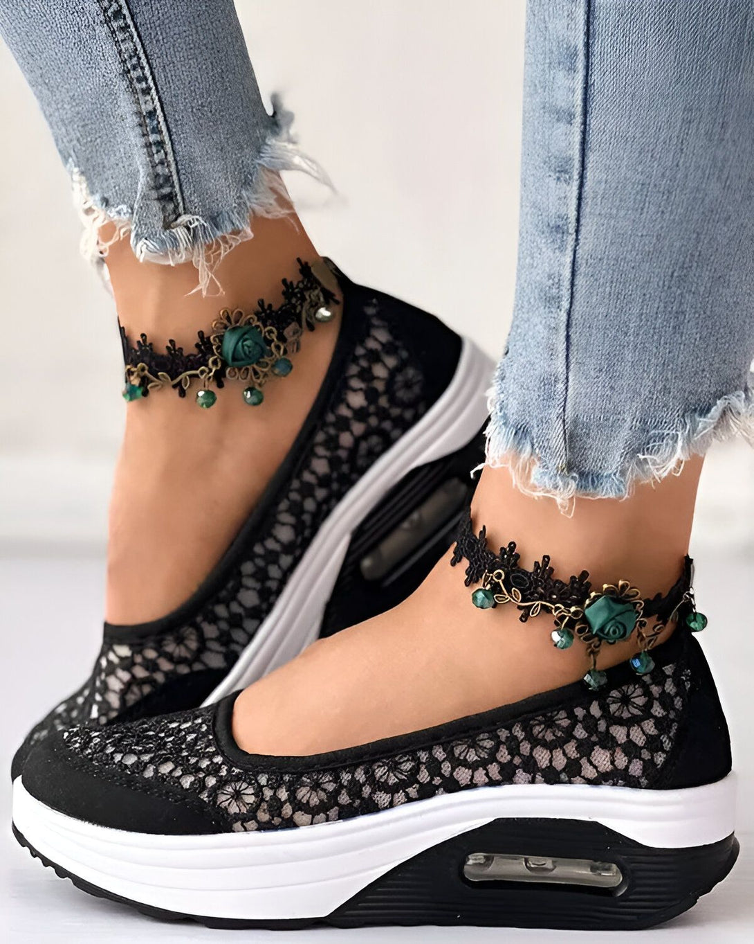 Une femme porte des chaussures noires en dentelle avec des semelles blanches. Les chaussures sont ornées de fleurs vertes et de perles. La femme porte également un jean bleu déchiré.