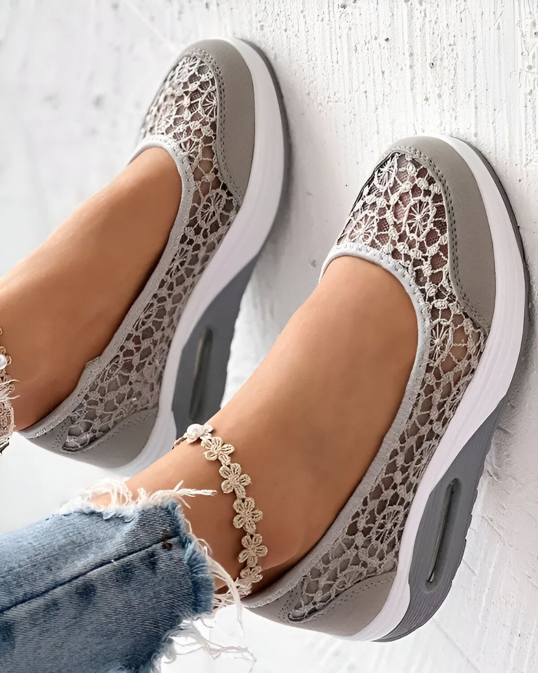 Une femme porte des chaussures de sport grises en dentelle avec une semelle blanche. Elle porte également un jean bleu et un bracelet de cheville en perles blanches.