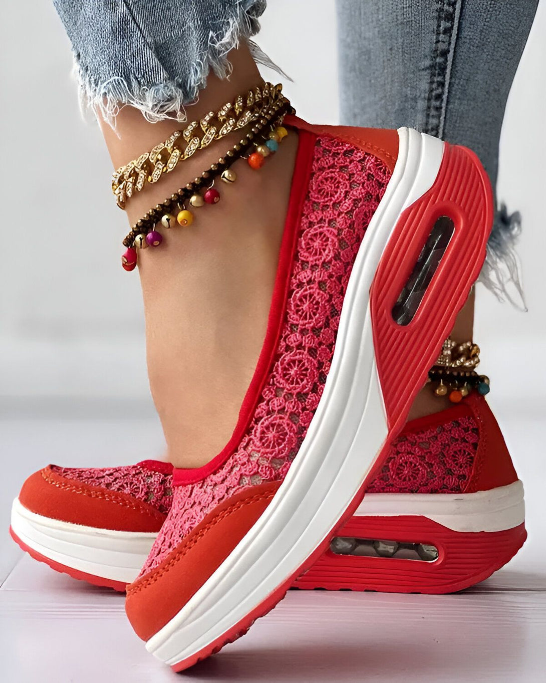 Une femme porte des baskets rouges en dentelle avec une semelle blanche. Elle porte également un jean bleu et des bijoux aux pieds.