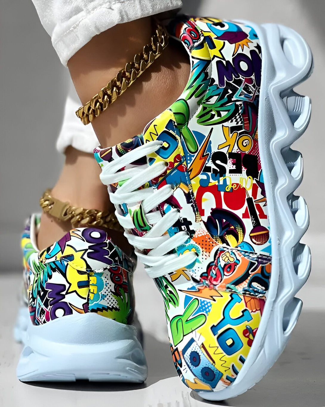 Une femme porte des baskets colorées avec des lacets blancs. Les baskets sont recouvertes de graffitis et de motifs colorés. La femme porte également un pantalon blanc et une chaîne en or à la cheville.