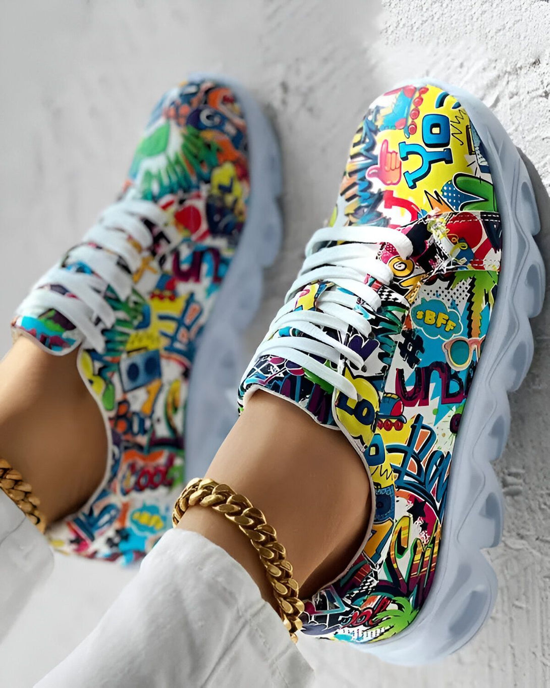 Une paire de baskets multicolores avec des lacets blancs et une semelle blanche. Les baskets sont portées par une femme qui porte un jean blanc et un t-shirt blanc. La femme est assise sur un mur blanc.