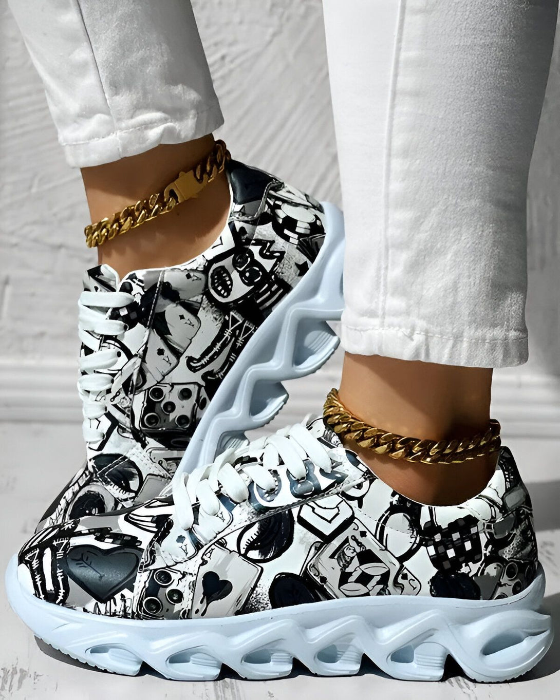 Une paire de baskets montantes à lacets blancs. Les baskets sont blanches avec un motif noir et gris. Il y a une chaîne en or autour de la cheville de la personne qui porte les baskets.