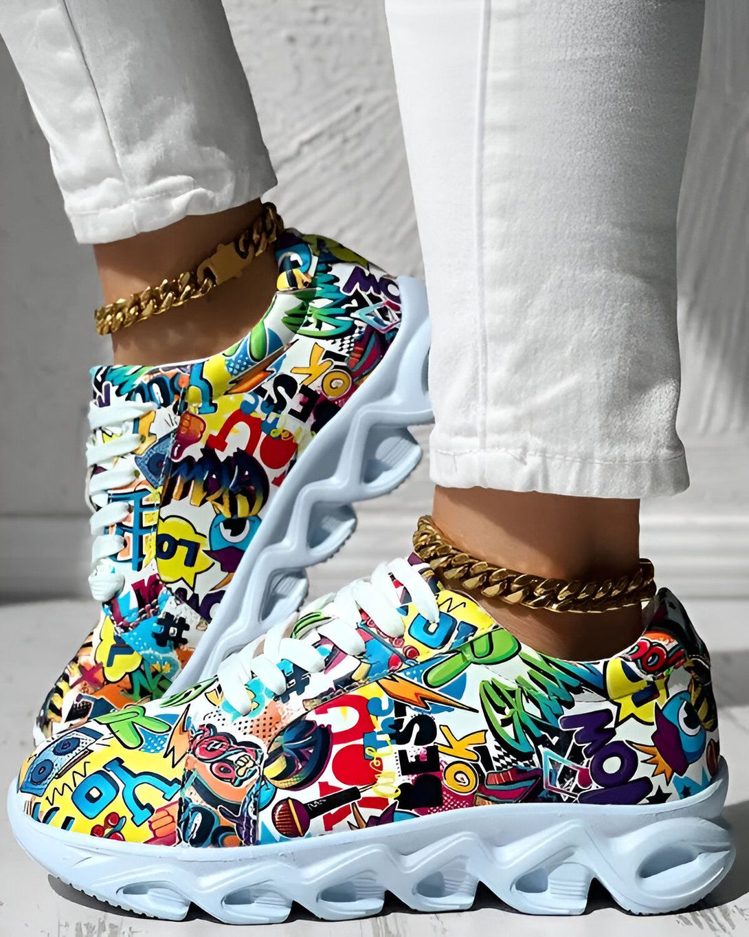 Une femme porte des baskets colorées avec un jean blanc et une chaîne en or à la cheville. Les baskets sont recouvertes de graffitis et ont des semelles épaisses.