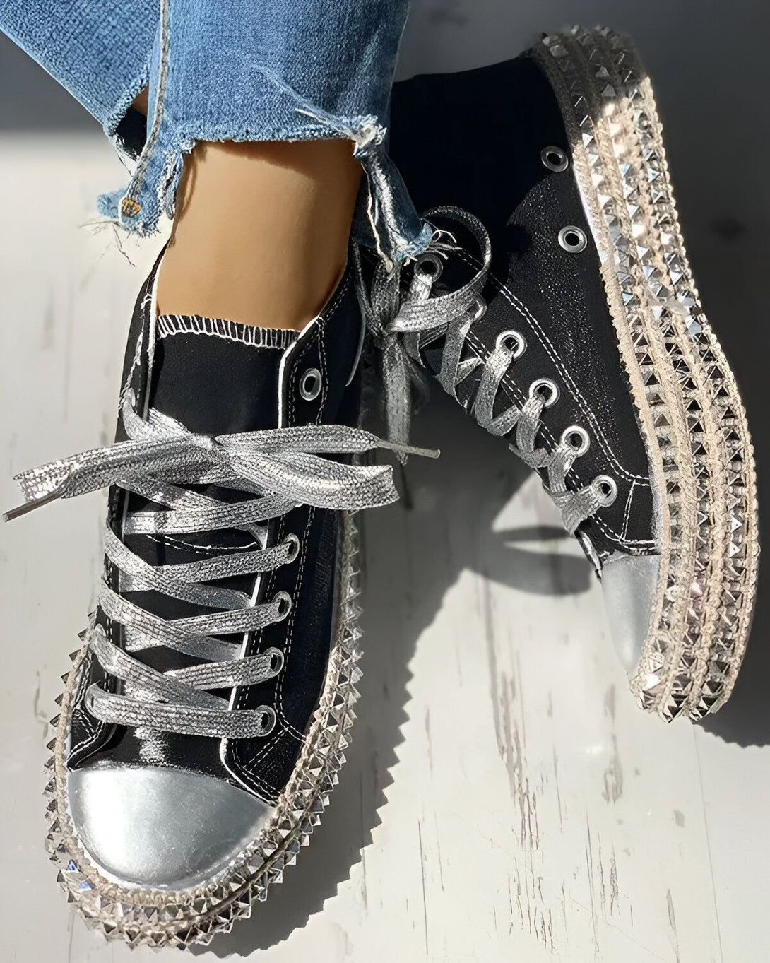 Une paire de sneakers noires à lacets argentés. Les sneakers sont cloutées sur le pourtour de la semelle.