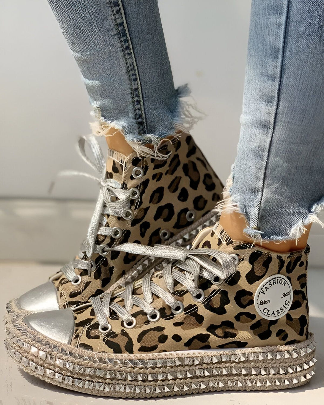 Une femme porte des baskets montantes à lacets. Les baskets sont en toile marron et imprimé léopard. Il y a des œillets et des lacets argentés. La semelle est en caoutchouc blanc avec des clous argentés. La femme porte un jean bleu troué.
