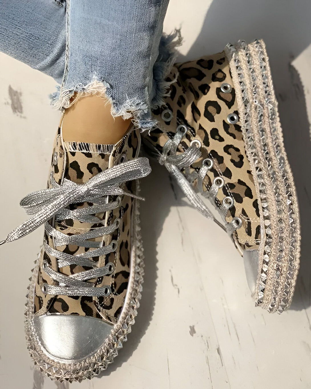 Une paire de sneakers montantes à lacets argentés. Les sneakers sont en toile marron et imprimé léopard, avec une semelle compensée blanche et des clous argentés sur le pourtour.