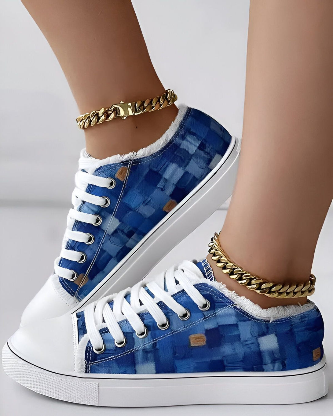 Une paire de baskets basses bleues et blanches avec des lacets blancs. Les baskets sont portées par une femme qui a une chaîne en or autour de la cheville.