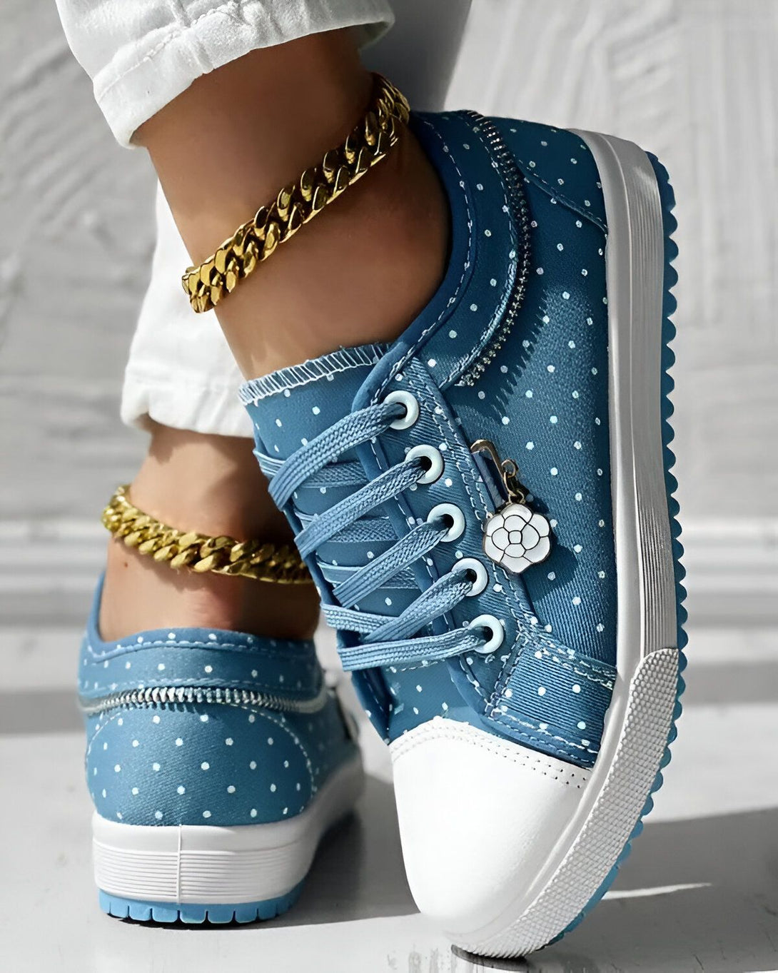 Une femme porte des baskets bleues à pois blancs. Les baskets sont lacées et ont une fermeture éclair sur le côté. La femme porte également un pantalon blanc et un bracelet de cheville doré.