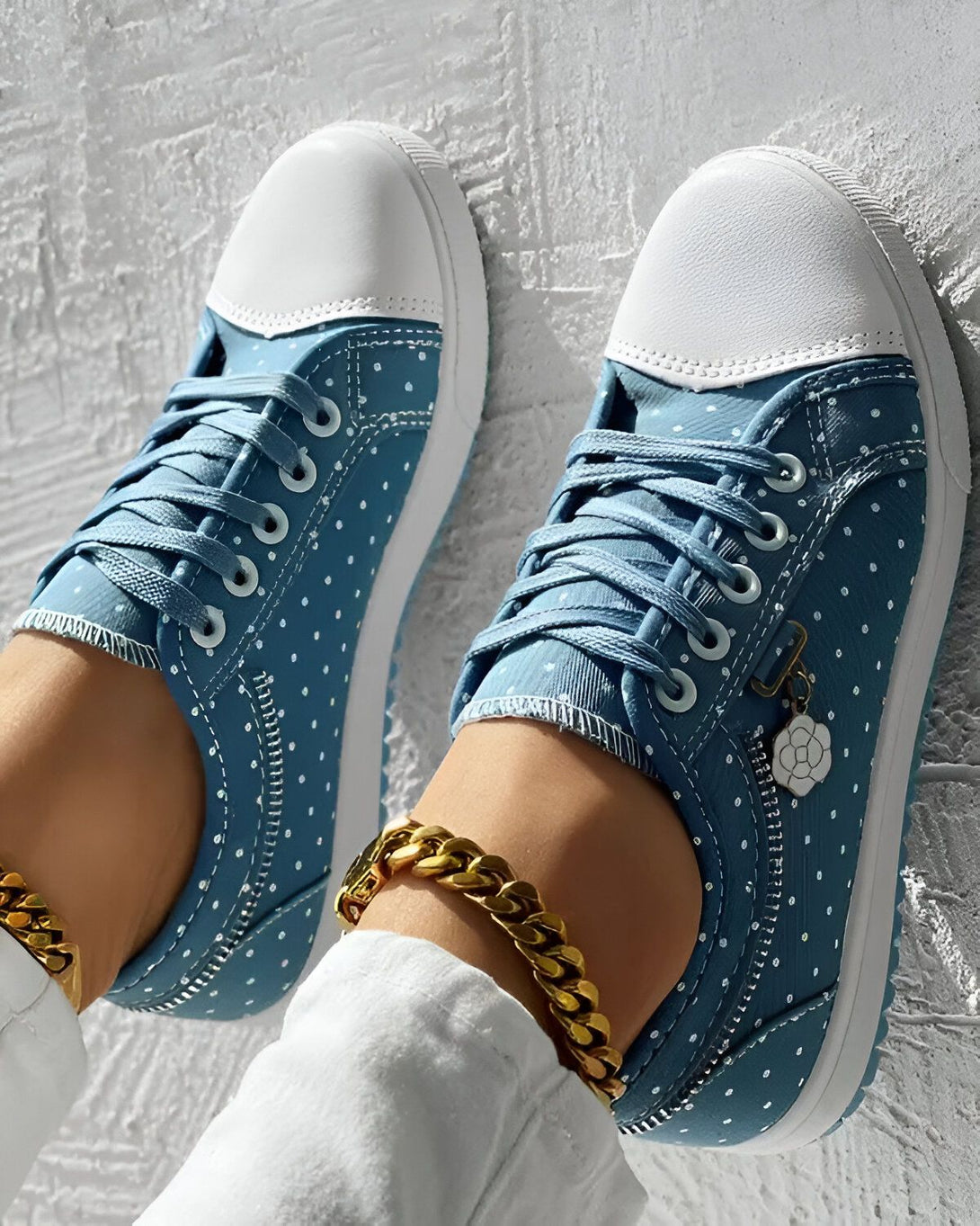 Une paire de chaussures bleues à pois blancs avec des lacets bleus et une semelle blanche. Il y a aussi un bracelet de cheville doré sur le pied gauche.