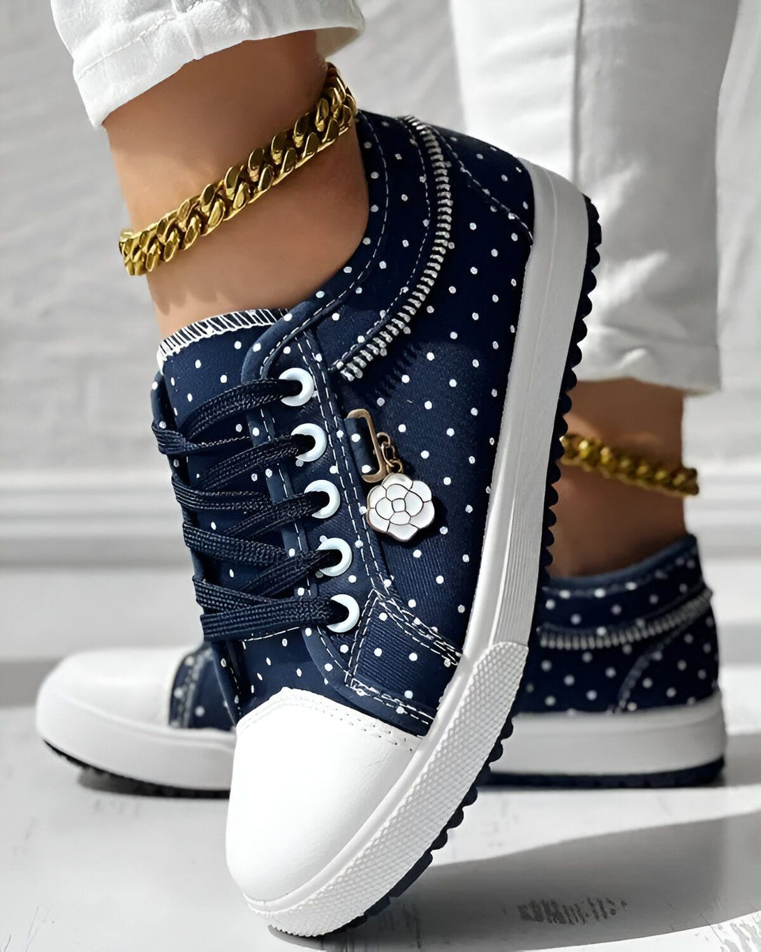 Une femme porte des baskets basses bleues à pois blancs. Les baskets sont lacées et ont une fermeture éclair sur le côté. La femme porte également un pantalon blanc et un bracelet de cheville doré.