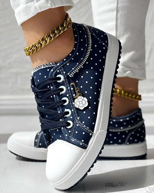 Une femme porte des baskets basses bleues à pois blancs. Les baskets sont lacées et ont une fermeture éclair sur le côté. La femme porte également un pantalon blanc et un bracelet de cheville doré.