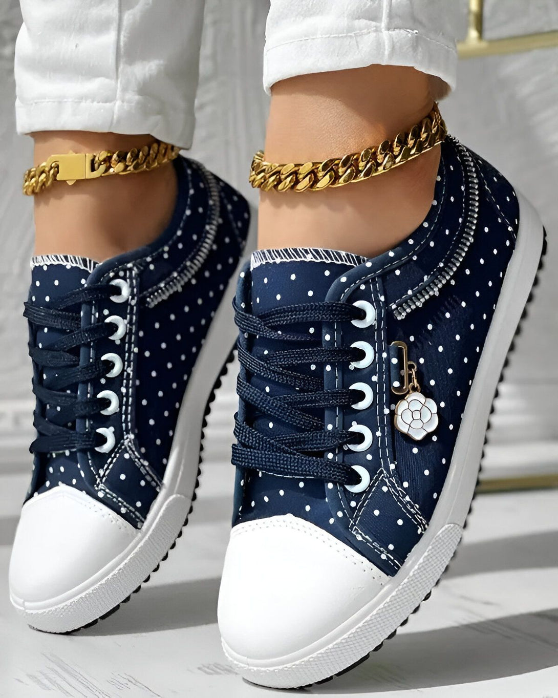 Une paire de chaussures basses bleues à pois blancs avec des lacets noirs et une semelle blanche. Les chaussures sont portées par une femme qui porte un pantalon blanc et un chemisier blanc.