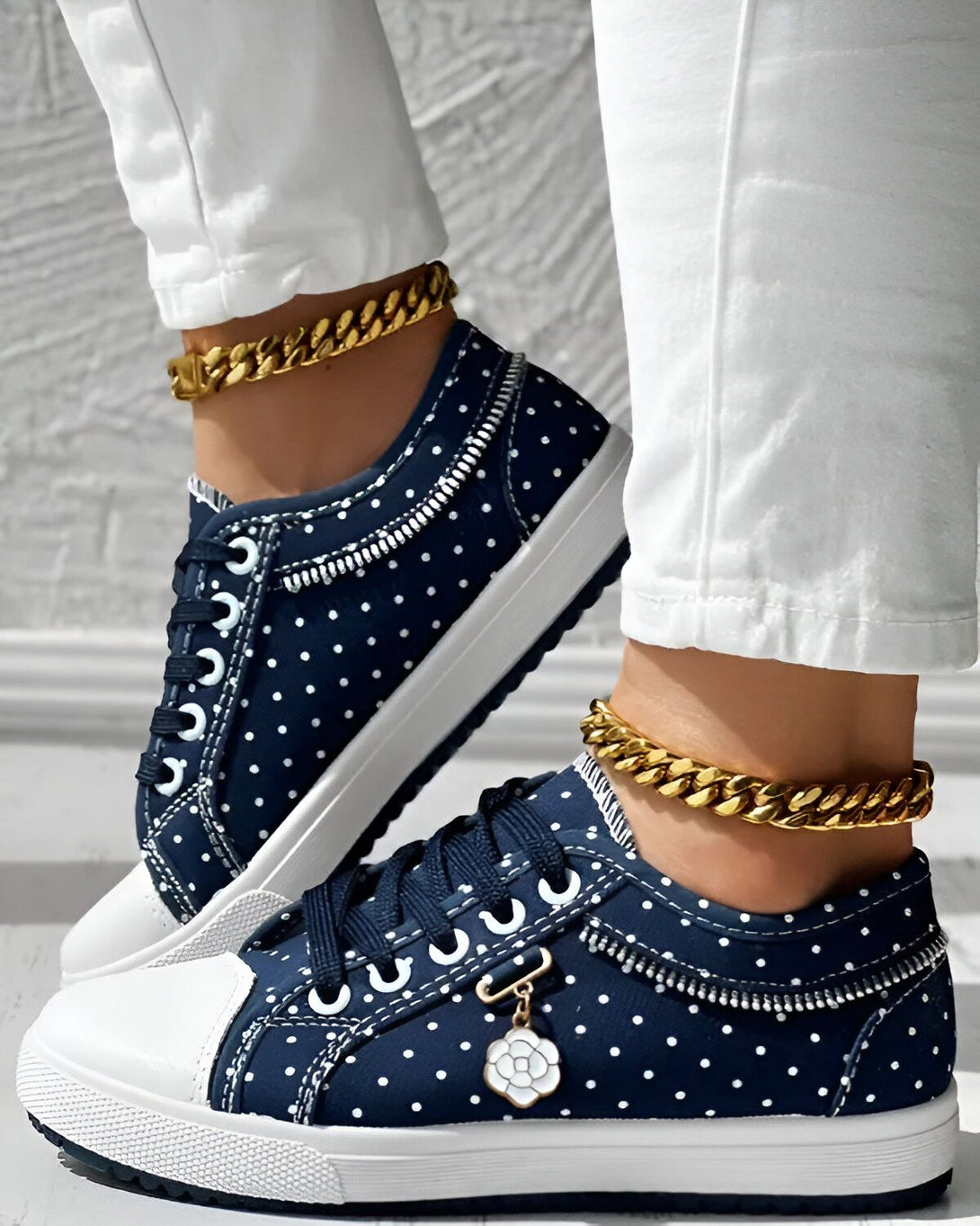 Une femme porte des chaussures en toile bleu marine à pois blancs. Les chaussures ont une semelle blanche et des lacets blancs. La femme porte également un pantalon blanc et des chaussettes blanches. Elle est debout sur un sol en béton.