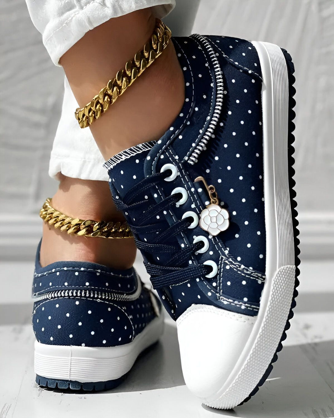 Une femme porte des baskets bleues à pois blancs. Les baskets sont lacées et ont une fermeture éclair sur le côté. La femme porte également un pantalon blanc et un bracelet de cheville doré.