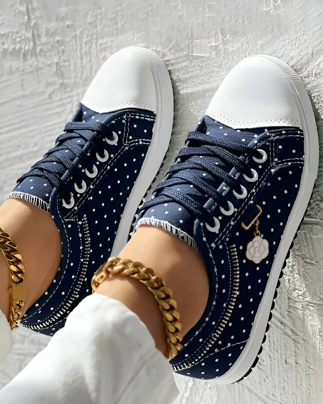 Une paire de chaussures bleues à pois blancs avec des lacets bleus et une fermeture éclair sur le côté. Il y a une chaîne en or avec un pendentif en forme de fleur blanche sur la cheville droite.