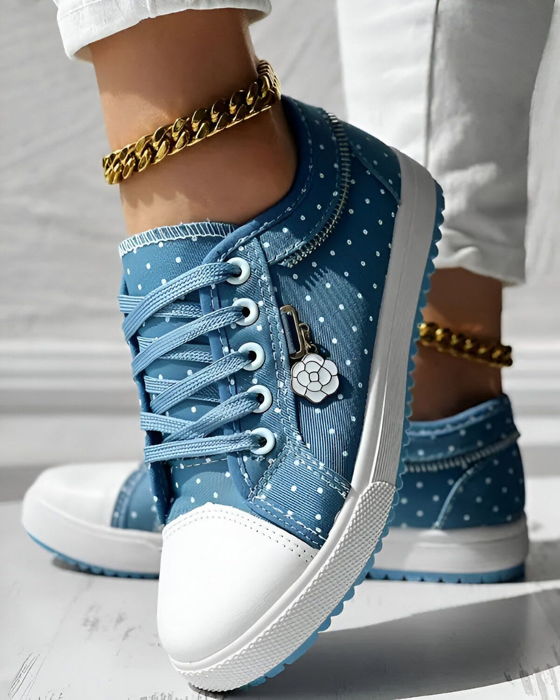 Une image d'une paire de chaussures bleues à pois blancs avec des lacets bleus et une semelle blanche. Il y a un charm en forme de fleur sur le côté de la chaussure. La personne porte également un bracelet de cheville en or.