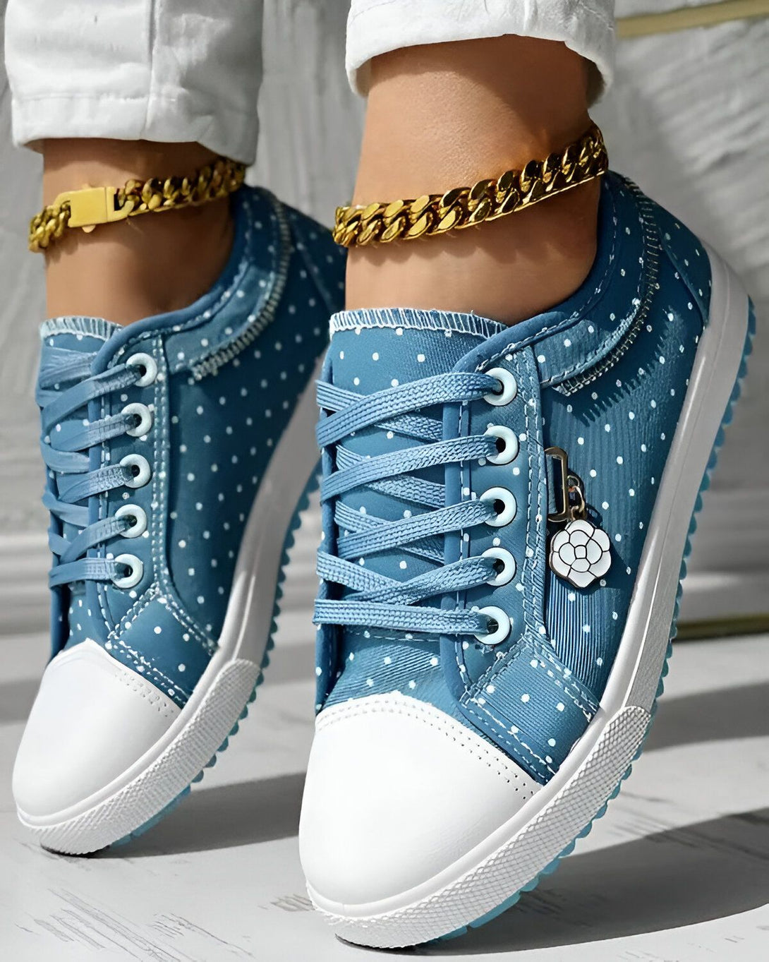 Une paire de chaussures en toile bleu clair à pois blancs, avec des lacets bleus et une semelle blanche. La chaussure est portée par une femme qui porte un pantalon blanc et un chemisier blanc. La femme est debout sur un sol en bois.