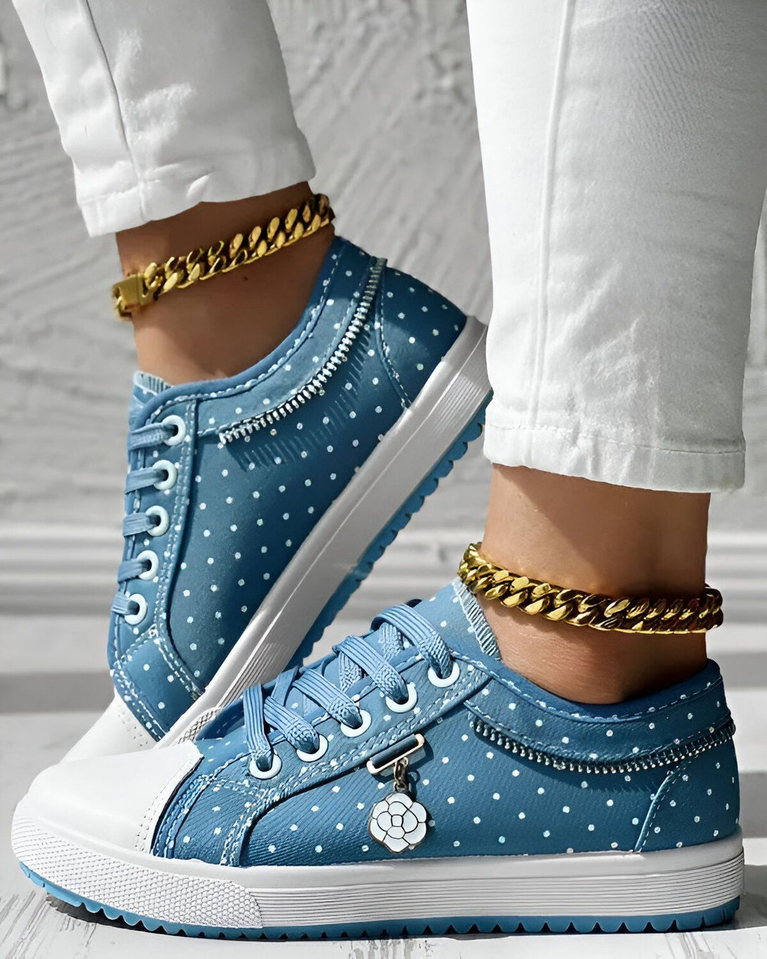 Une femme porte des baskets bleues à pois blancs. Les baskets ont des lacets blancs et une semelle blanche. La femme porte également un jean blanc et des chaussettes blanches. Elle est debout sur un sol en bois.