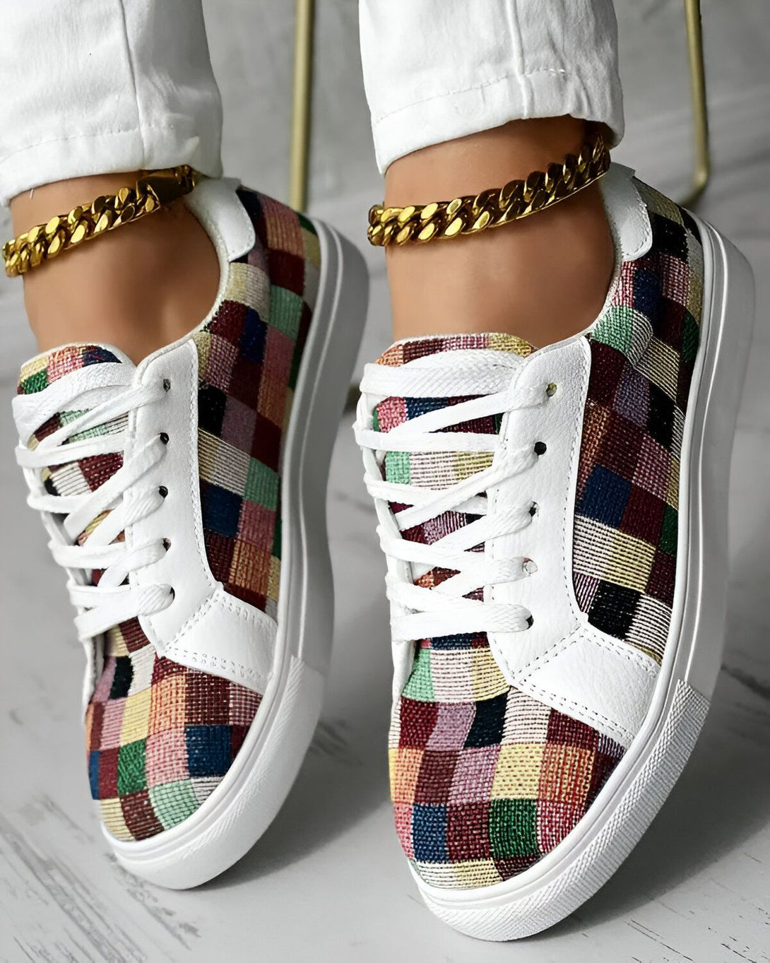 Une femme porte des baskets basses à lacets avec un motif à carreaux multicolores. Les baskets ont une semelle blanche et des lacets blancs. La femme porte également un pantalon blanc et des chaussettes basses blanches.