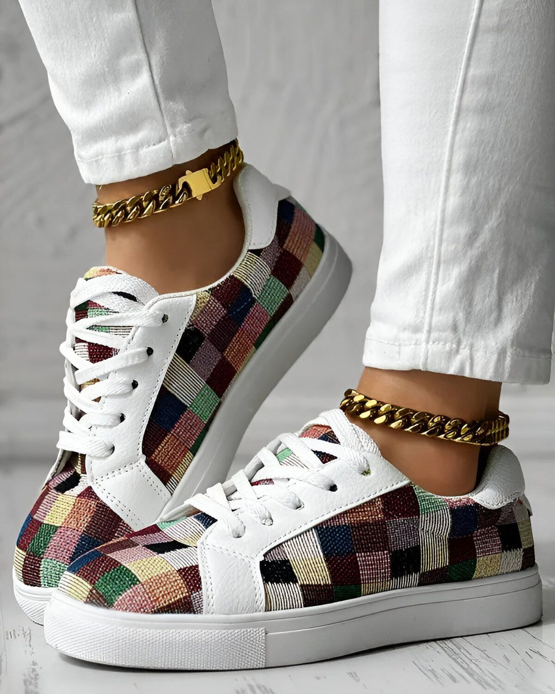 Une femme porte des baskets basses à lacets avec un motif à carreaux multicolores. Elle porte également un jean blanc et des bracelets de cheville en chaîne dorée.