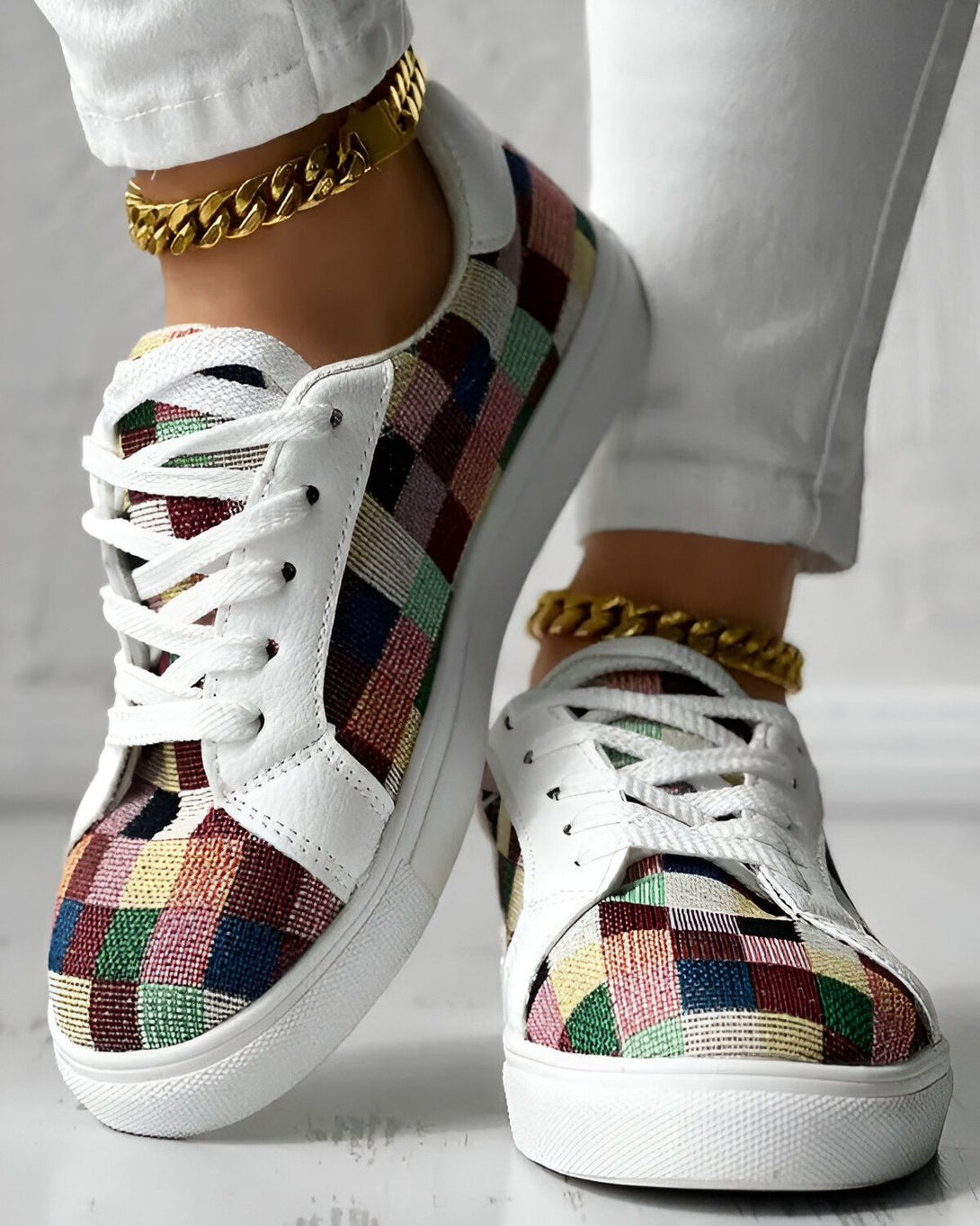 Une femme porte des baskets basses à lacets avec un motif à carreaux multicolores. Les baskets ont une semelle blanche et des lacets blancs. La femme porte également un jean blanc et des bijoux en or.