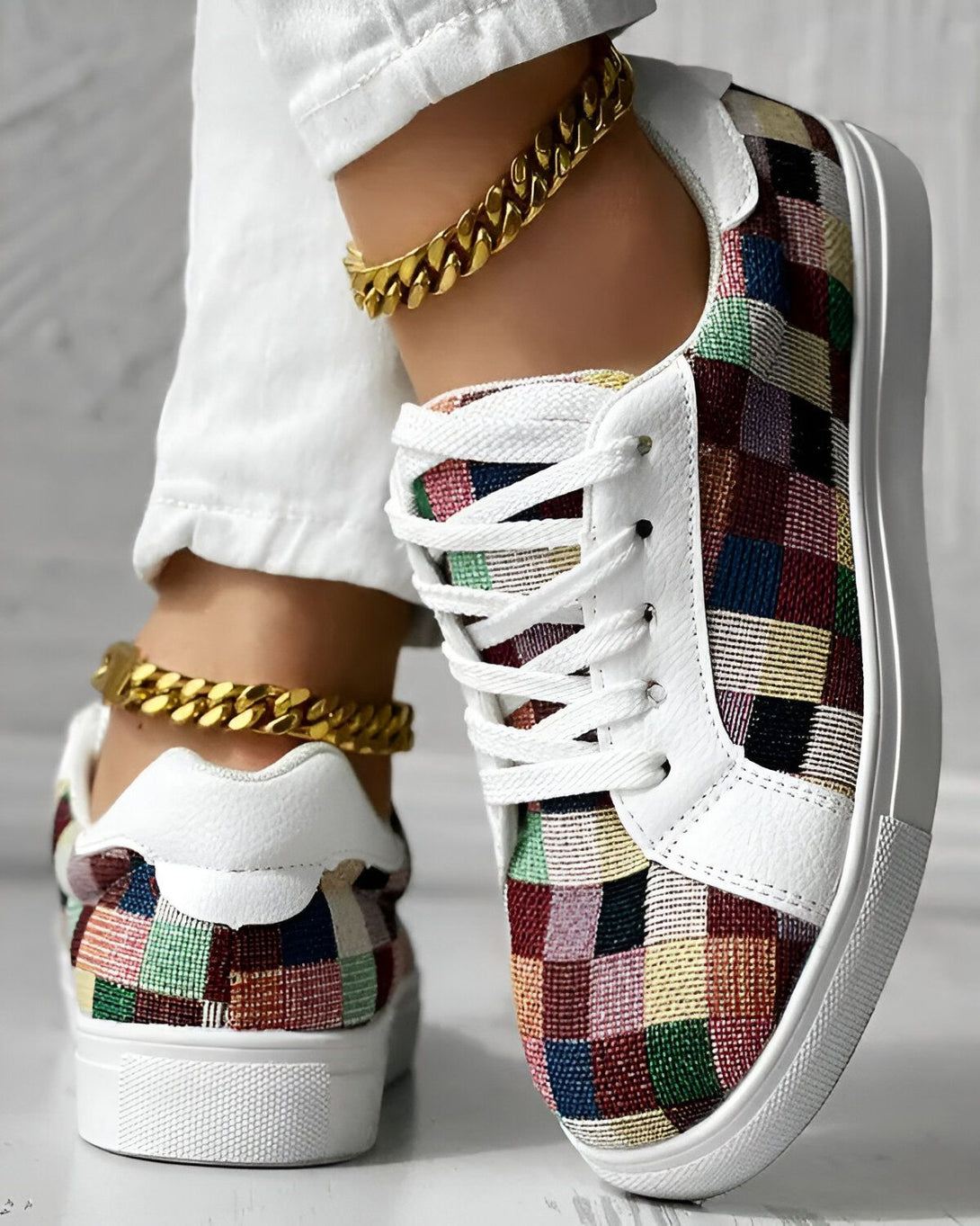 Une femme porte des baskets colorées avec un pantalon blanc et des chaussettes basses. Les baskets sont en toile et ont une semelle en caoutchouc blanche. La femme a une chaîne en or autour de la cheville.